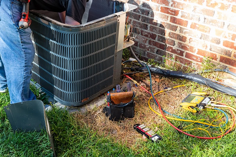 3 Tips to Prevent AC Breakdown During a Heat Wave.  Photo of an air conditioning unit being worked on by a technician outside of a brick home.