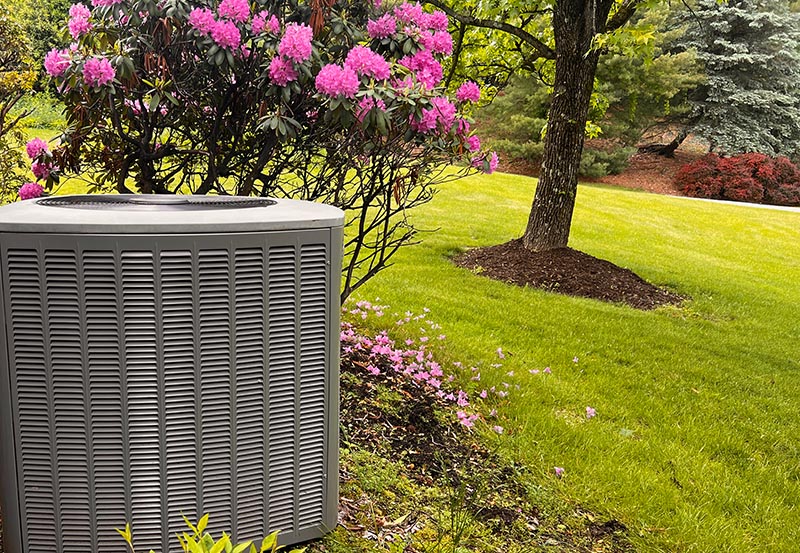 An outdoor central air conditioning condenser or heat pump unit situated in a beautifully landscaped yard.