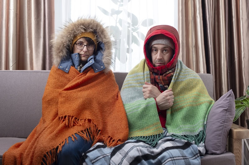 A man and a woman sitting on a couch indoors, looking uncomfortably cold.
